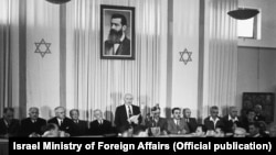 Prvi premijer Izraela David Ben-Gurion (stoji ispod portreta Teodora Hercla, osnivača i lidera cionističkog pokreta), okružen članovima Nacionalnog jevrejskog saveta, u 18.01 časova 14. maja 1948. u Tel Avivu zvanično proglašava državu Izrael.