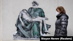 Egy járókelő sétál el egy falra festett kép előtt, amely egy orvost ábrázol Michelangelo Pietájára utaló pózban, egy kórház közelében, Szófiában. 