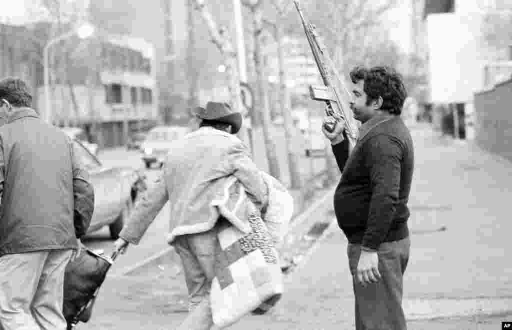 A U.S.citizen carries belongings past an armed Khomeini loyalist who is guarding an evacuee staging area in Tehran on February 17, 1979. Hundreds of Americans began to flee Iran following the first attempt to seize the U.S. Embassy.