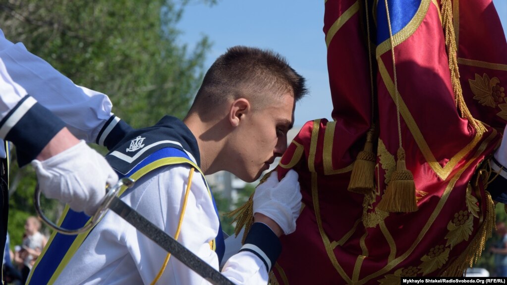 Старий прапороносець цілує прапор на прощання і передає його молодому, який також торкається губами знамені
