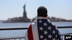 Иммигрант, накинувший на себя флаг США, смотрит на Статую Свободы, расположенной на острове Свободы в примерно в 3 км на юго-запад от южной оконечности Манхэттена, в штате Нью-Йорк. 