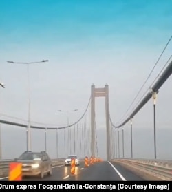 În depărtare se văd pilonii înalți, de beton, care asigură, alături de blocurile în care sunt ancorați, menținerea tensiunii cablurilor podului suspendat.