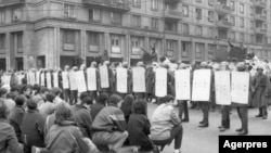 Revoluția Română, 21 Decembrie 1989, Piața Romană din București. Manifestanții se așezaseră în genunchi în fața cordonului de militari, în spatele cărora erau tancuri. Manifestanții arătau că sunt pașnici. În cursul nopții, forțele de armată și securitate vor deschide focul.
