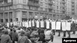 Barajul de scutieri, întărit de tancuri, din Piața Romană, în fața unui grup de manifestanți care îngenunchează. Gestul e frecvent în multe din episoadele Revoluției. Era un gest de protest față de ateismul militant al comunismului, dar și o formă de a arăta că ei, demonstranții, resping violența.