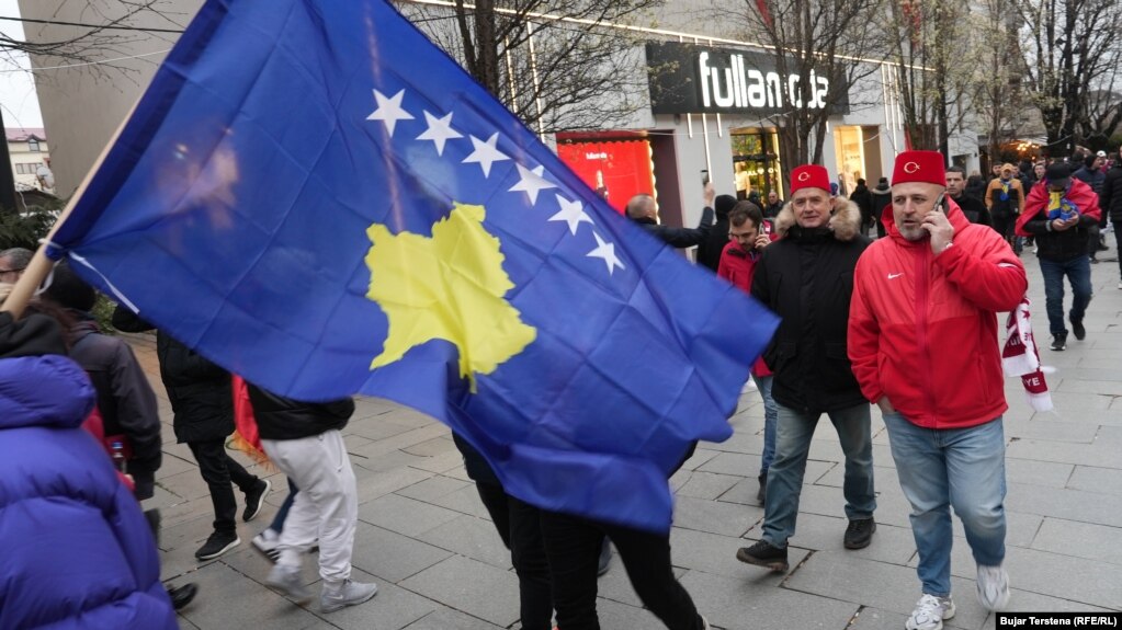 Dy tifozë tuq shihen duke ecur në sheshin e Prishtinës mbrapa një flamuri të Kosovës, disa orë para ndeshjes vendimtare mes Kosovës dhe Turqisë për një vend në Kupën e Botës 2026, të martën në Stadiumin "Fadil Vokrri".