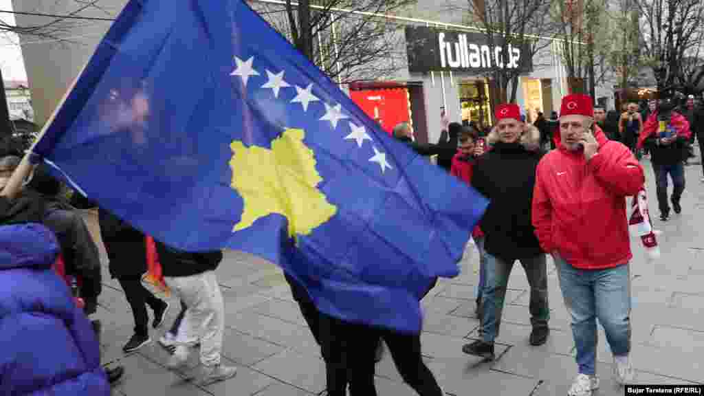 Dy tifozë tuq shihen duke ecur në sheshin e Prishtinës mbrapa një flamuri të Kosovës, disa orë para ndeshjes vendimtare mes Kosovës dhe Turqisë për një vend në Kupën e Botës 2026, të martën në Stadiumin "Fadil Vokrri".