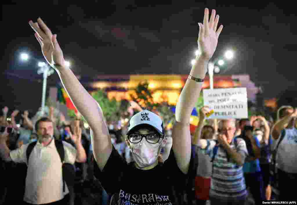 Oamenii ridică mâinile în timp ce poartă măști în timpul unui protest în fața sediului guvernului român din București la 17 august 2018.&nbsp; Sute de protestatari s-au adunat în Piața Victoriei pentru a marca o săptămână de la violențele Jandarmeriei asupra populației pașnice, împotriva căreia a folosit tunuri cu apă, gaze lacrimogene, spray cu piper și bastoane, fugărind oamenii pe străzile adiacente și snopindu-i în bătaie. Peste 400 de persoane au fost rănite și 30 arestate.&nbsp;