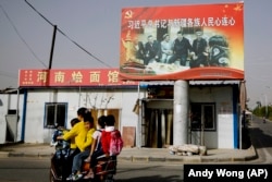 A Uyghur woman in Xinjiang on a scooter with schoolchildren as they ride past a picture showing Chinese leader Xi Jinping joining hands with a group of Uyghur elders.