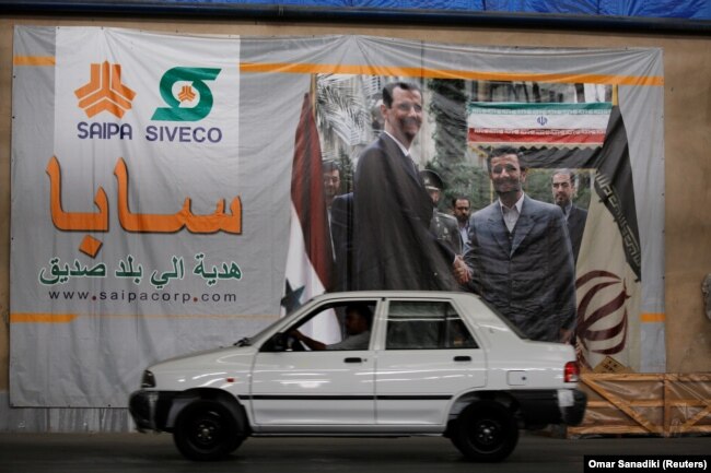 یک خودرو سایپا در حال عبور از مقابل تصویر بشار اسد و محمود احمدی‌نژاد در حمص سوریه/ این تصویر در شهریور ۹۷ گرفته شده است