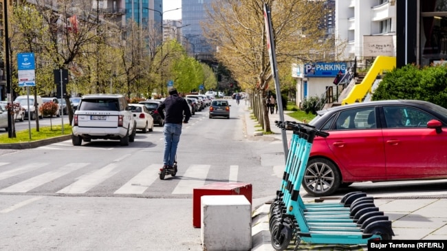 Një person duke udhëtuar nëpër rrugët e Prishtinës me një trotinet, teksa në anën e djathtë të fotografisë shihen disa trotinetë elektrikë, që qytetarët mund t'i përdorin, në këmbim të një pagese.