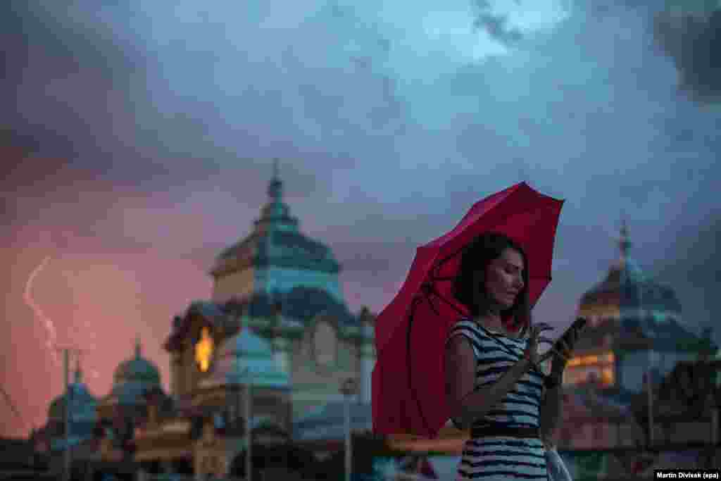 Një grua me ombrellë derisa përdor telefonin e saj celular, ndërkaq në sfond shihet rrufeja, Pragë, Republika Çeke.