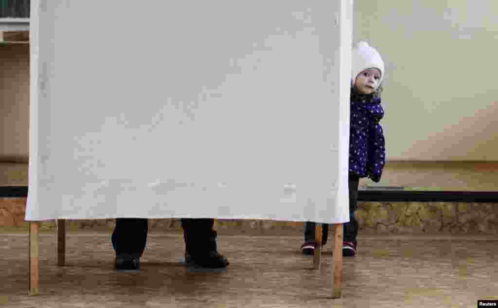 A child looks out from behind a curtain as her mother fills up her ballot.