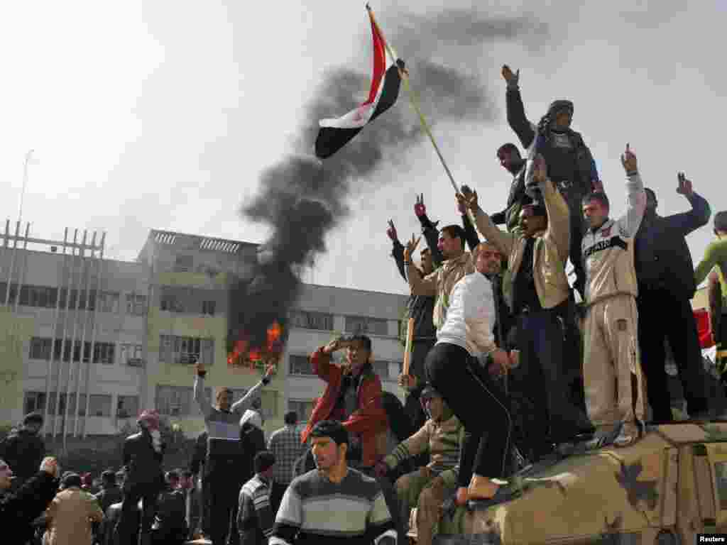 Protesters atop a military vehicle as smoke rises from the Mosul governorate building.