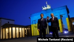 9 mai 2022, la Berlin: președintele francez Emmanuel Macron și cancelarul german Olaf Scholz în fața Porții Brandenburg, iliuminată în culorile naționale ale Ucrainei, în semn de solidaritate cu țara atacată de Rusia. 