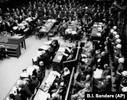 Засідання Нюрнберзького процесу, 20 листопада 1945 року. Військова поліція стоїть у залі суду. На передньому плані сидять підсудні, а перед ними сидять їхні захисники