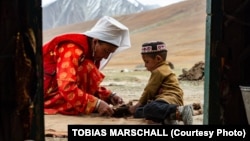 A Pamir Kyrgyz mother cuts the fabric of a rug to stitch together new clothes, studied closely by her son. 