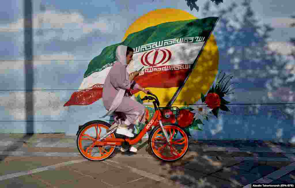Іранская жанчына на ровары ў Тэгеране на тле графіці з нацыянальным сьцягам Ірану.