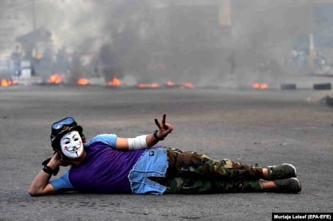 An Iraqi protester wearing a Guy Fawkes mask flashes victory signs in front a fire set by protesters to block a street during a protest and strike at Khillani square in central Baghdad, November 4, 2019