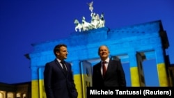 Канцлер Германии Олаф Шольц (справа) и президент Франции Эмманюэль Макрон у Бранденбургских ворот, подсвеченных в цвета украинского флага, во время масштабного вторжения России в Украину. Берлин, 9 мая 2022 года