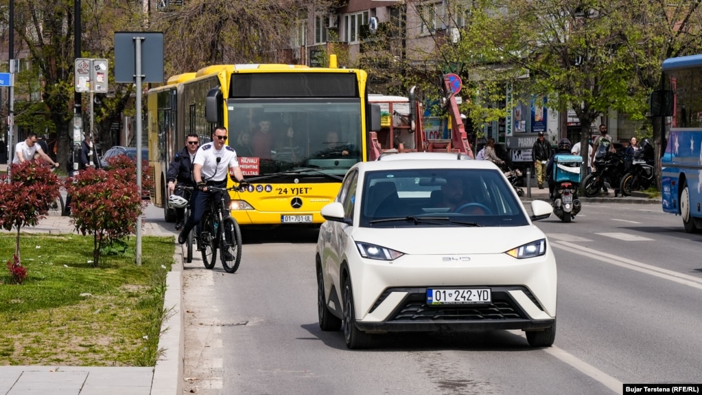 Një veturë elektrike shihet në rrugët e Prishtinës, teksa pas tij është një autobus i një linje private që operon në Prishtinë. Për të nxitur qytetarët që të përdorin transportin publik, në Prishtinë, autoritetet lokale kanë bërë integrimin e biletave të autobusëve, duke u mundësuar qytetarëve që me një biletë (bileta ditore kushton 0.80 centë) të mund të udhëtojnë me të gjitha linjat brenda një dite.