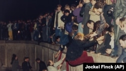 Fotografie din 10 noiembrie 1989 îi arată pe berlinezii de est primind ajutor de la berlinezii de vest în timp ce escaladează Zidul Berlinului care a împărțit orașul de la sfârșitul celui de-al Doilea Război Mondial, lângă Poarta Brandenburg.