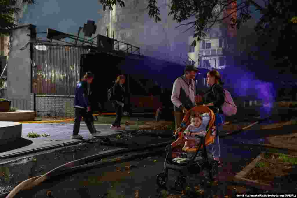 Родина з візочком чекає на вулиці, доки загасять пожежу в їхньому будинку, який зайнявся внаслідок російського обстрілу, Київ, 19 липня 2026 року