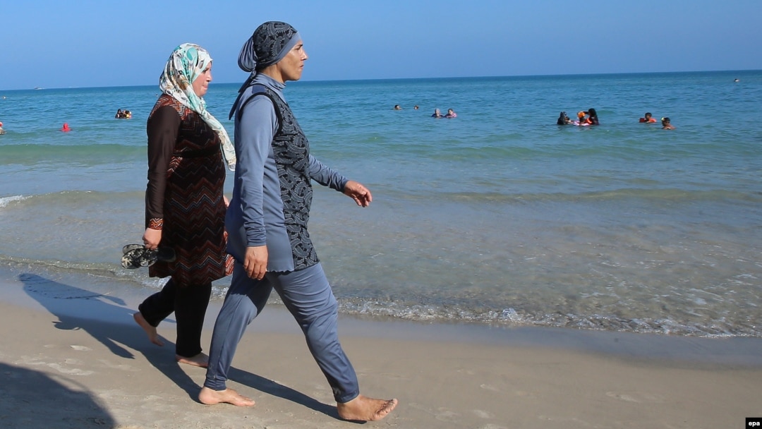 Women walk on a beach wearing burkinis.