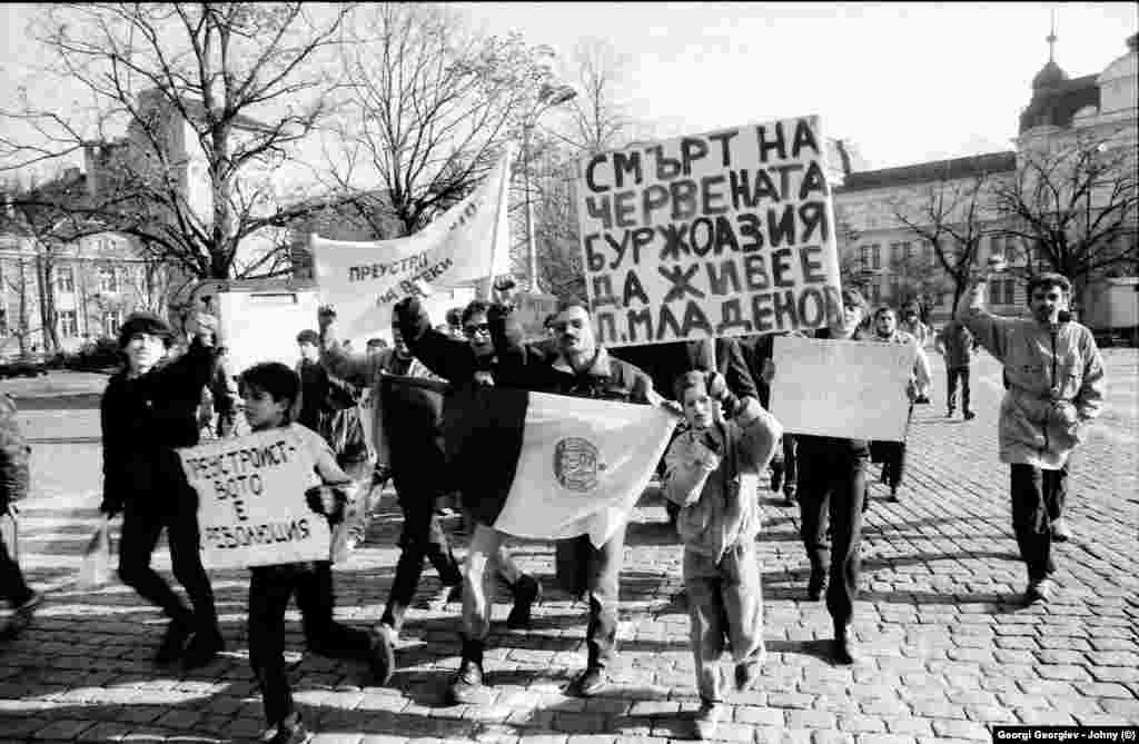 Група хора са се запътили към митинга на опозицията от 18.11.1989 г. с плакат, героизиращ Петър Младенов - член на Политбюро и външен министър на Тодор Живков. Самият митинг гласува &quot;вот на доверие&quot; на Петър Младенов, оглавил БКП след свалянето на диктатора. В онези дни, когато още не е ясно дали системата трябва да се облагородява или да бъде изтръгната с корен, Младенов е героят както на комунистите, така и на не-комунистите. Той върви по улицата без охрана.