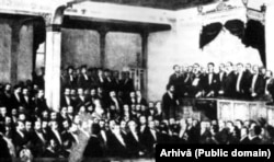 Carol I de Hohenzollern Sigmaringen depune jurământul la 10 Mai 1866, în fața Adunării Naționale de la București. A intrat incognito în România, din cauza războiului dintre Prusia și Austria, dar a reușit să impiedice pe muche de cuțit reocuparea Principatelor de către armatele otomane și rusești. Litografie, Arhivele Naționale.