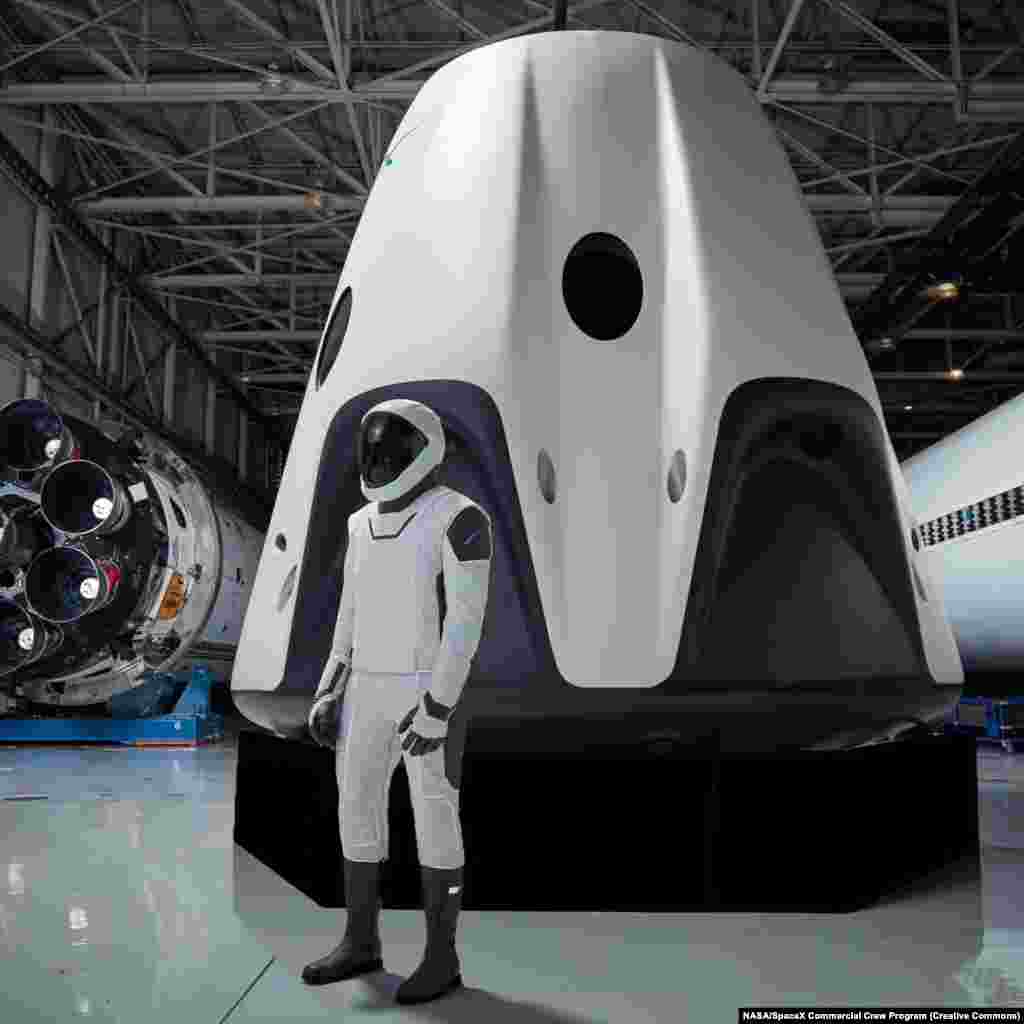 Noul modul SpaceX Crew Dragon. Costumul spațial SpaceX care va fi purtat de astronauți la bordul navetei spațiale Crew Dragon (în fundal) în timpul misiunilor către și dinspre Stația Spațială Internațională. SpaceX își dezvoltă naveta spațială Crew Dragon și racheta Falcon 9 în parteneriat cu Programul comercial de echipaj al NASA pentru a transporta astronauți de la și dinspre stația spațială.