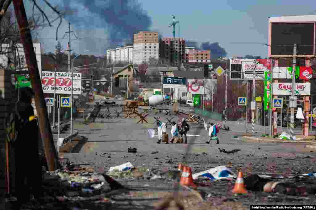 Цивільні з білою тканиною поверх одягу виходять з Бучі. Вулиця Соборна, що розділяє Бучу і Ірпінь. 11 березня 2022 року.Кордон між смертю і життям, на горизонті – окупована Буча. Жителі покидають місто. З сусіднього супермаркету чоловік виносить спортивний лук, його кличе до себе військовий на крайньому блокпосту, забирає лук і відправляє чоловіка до поліції для зʼясування особи.«Звідти ось так виходять, всі в білому. А тих, хто хоче зайти назад у Бучу, росіяни вбивають», – каже військовий