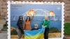 Жанчыны з украінскім нацыянальным сьцягам побач з арт-аб'ектам з выява новай маркі з Керчанскім мостам, які палае. Ілюстрацыйнае фота