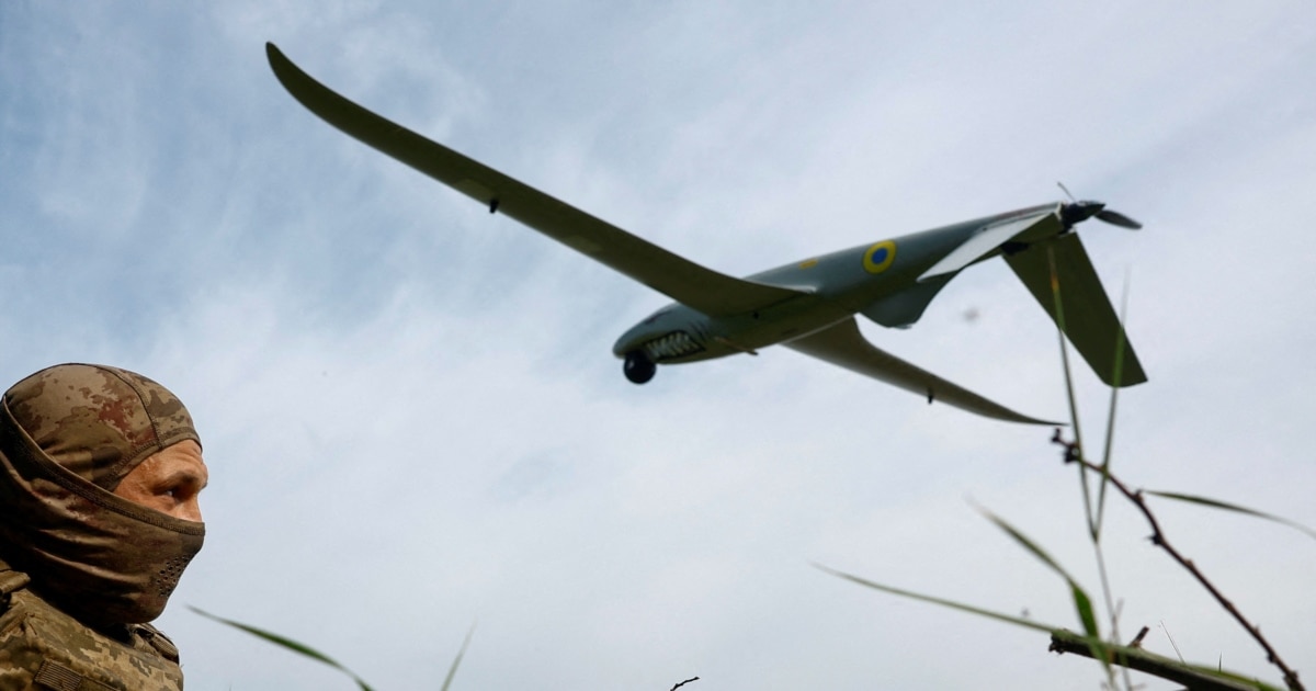 A person in camouflage watches a small blue and yellow aircraft flying overhead.