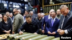 Ukrainian President Volodymyr Zelenskiy (signing a shell in the center of the picture) visits a munitions manufacturing plant with Ukrainian Ambassador to the United States Oksana Markarova in the background (far left). 