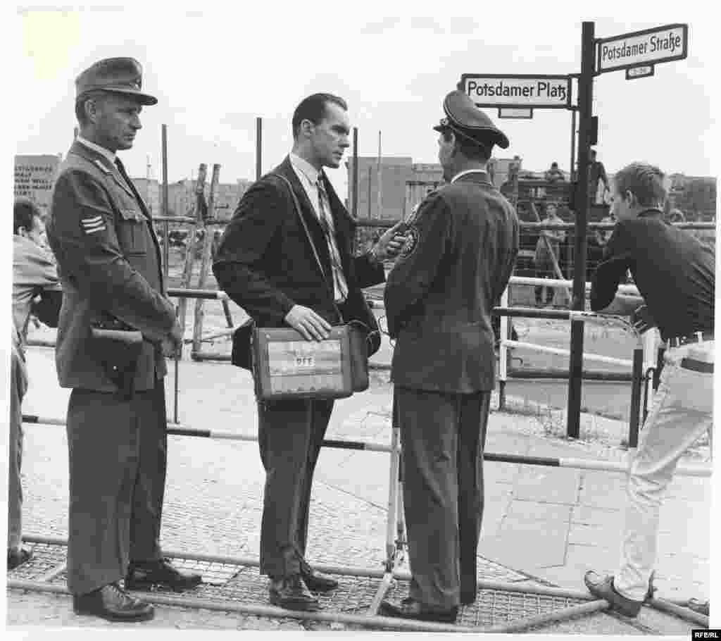 William Marsh, Berlin bureau chief of Radio Free Europe, interviews a police official at Potsdamer Platz. - In the background, East German workers are erecting the permanent wall, which bisected Potsdamer Platz and rendered the square a desolate wasteland.