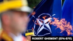 Un soldat român din garda de onoare, susținut de drapelul NATO, stă nemișcat în timpul ceremoniei de depunere a coroanei, așa cum se vede prin flacăra eternă, la Memorialul Soldatului Necunoscut, cu ocazia comemorării Zilei Eroilor, la București, România, 28 mai 2020.