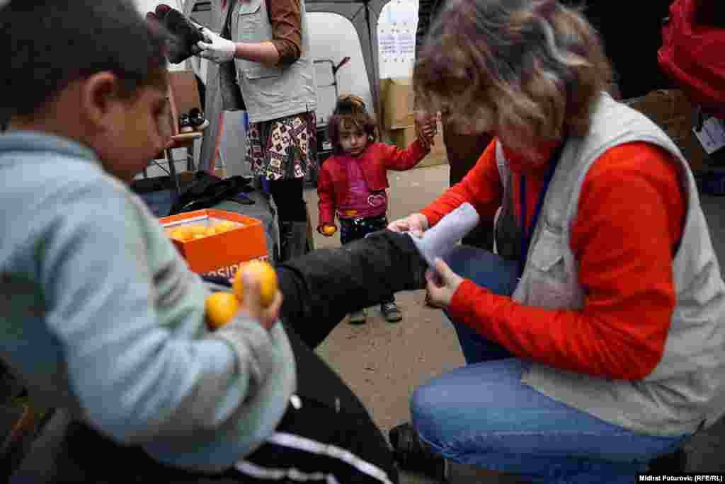 Djevojčica gleda kako član humanitarne organizacije dječaku oblači čarape u prihvatnom centru u Opatovcu, pored grada Tovarnik u Hrvatskoj.Ovdje se vrši registracija izbjeglica koje su iz Srbije došle u Hrvatsku. Nakon registracije, izbjeglice nastavljaju put prema Sloveniji, većina izbjeglica želi da stigne u Njemačku.