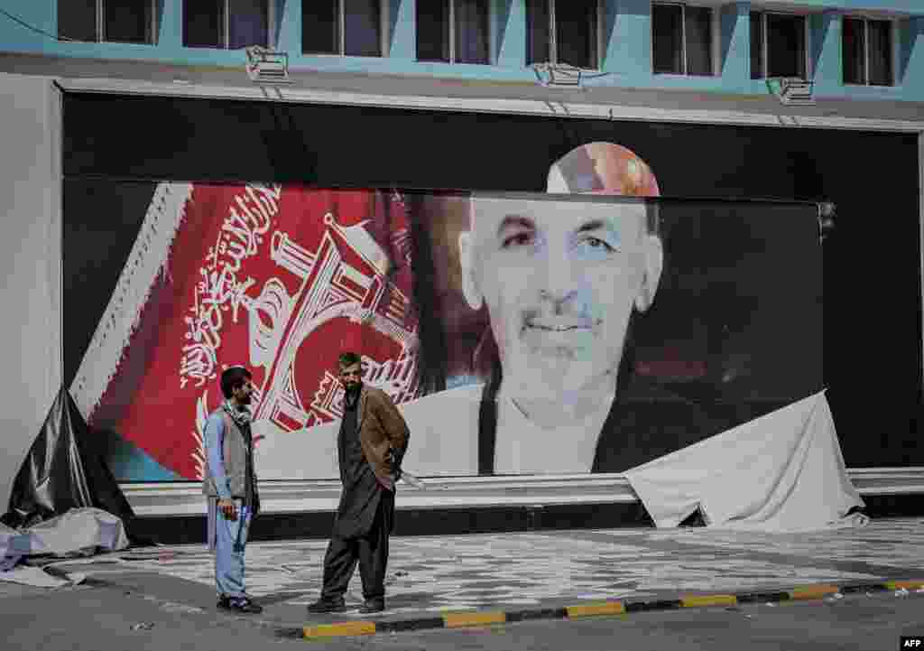 Афганистанци разговарят до постер с лика на избягалия афганистански президент Ашраф Гани на летището в Кабул.
