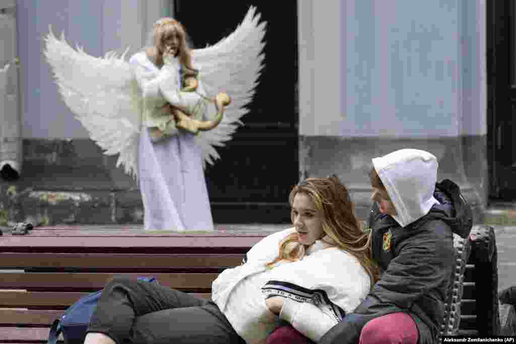 Молодая пара отдыхает на скамейке, пока уличная актриса в костюме ангела разговаривает по телефону на Никольской улице возле Кремля в Москве