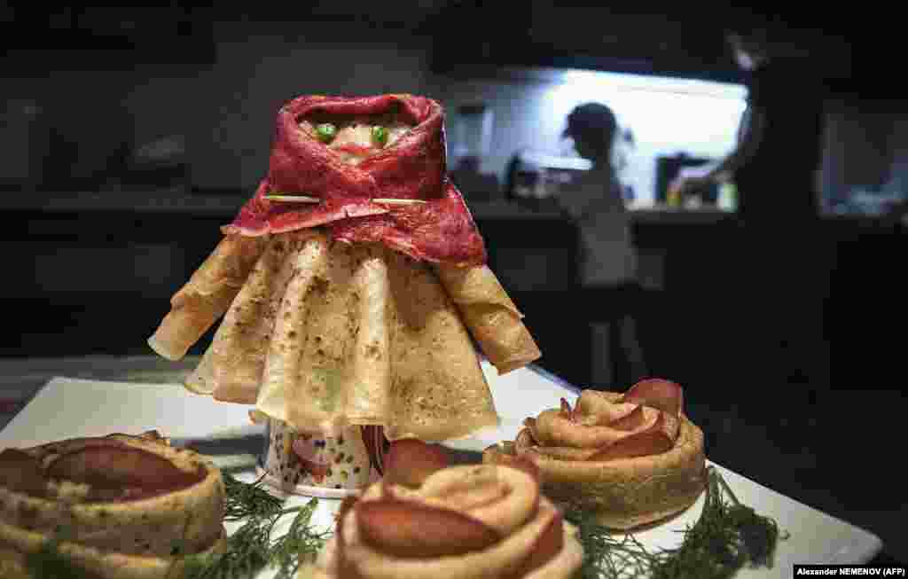 A family cooks a holiday dinner at their apartment in Moscow as a pancake woman stands on a table.