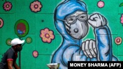 Egy gyümölcsárus húzza a szekerét egy falfestmény mellett, amely a járvány elleni védekezés frontvonalában dolgozó maszkos embert ábrázol Újdelhiben, 2021. június 27-én