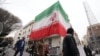 TOPSHOT - A huge Iranian flag adorns a building as Iranians walk past damaged structures following an earlier military strike in the Iranian capital, Tehran, on March 15, 2026.
