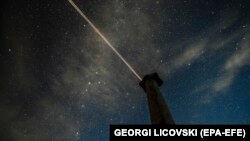 Repülőgép húz maga után csíkot az égen, miközben elvonulnak a Perseidák Észak-Macedónia római kori romjai, a Stobi felett augusztus 12-én.