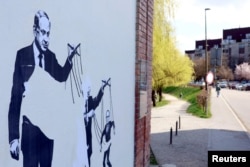 Një mural që shfaq kryeministrin Izraelit Netanyahu duke e kontrolluar ish-kryeministrin slloven, Janez Jansha, i cili po ashtu sfaqet duke tërhequr fijet e figurës së Anzhe Llogarit.