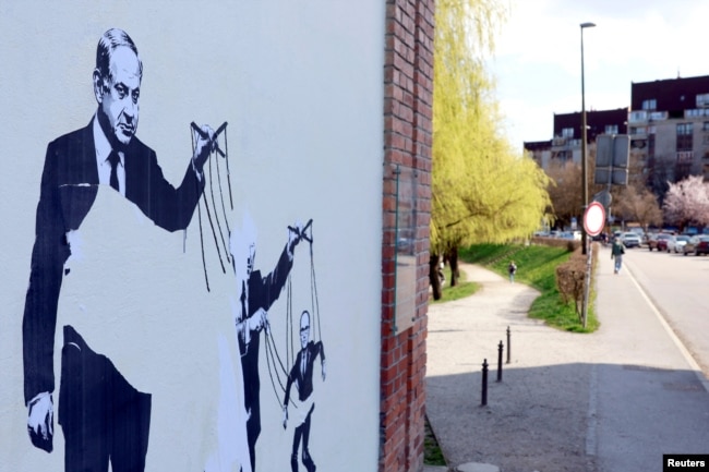 Një mural që shfaq kryeministrin Izraelit Netanyahu duke e kontrolluar ish-kryeministrin slloven, Janez Jansha, i cili po ashtu sfaqet duke tërhequr fijet e figurës së Anzhe Llogarit.