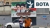 Dy gra ecin në një rrugë në Teheran para një pankarte me portrete të tre burrave që kanë qenë udhëheqës supremë të Republikës Islamike: ajatollah Ruhollah Khomeini (majtas), ajatollah Ali Khamenei (në qendër) dhe lideri i tanishëm, Mojtaba Khamenei.