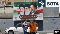 Dy gra ecin në një rrugë në Teheran para një pankarte me portrete të tre burrave që kanë qenë udhëheqës supremë të Republikës Islamike: ajatollah Ruhollah Khomeini (majtas), ajatollah Ali Khamenei (në qendër) dhe lideri i tanishëm, Mojtaba Khamenei.