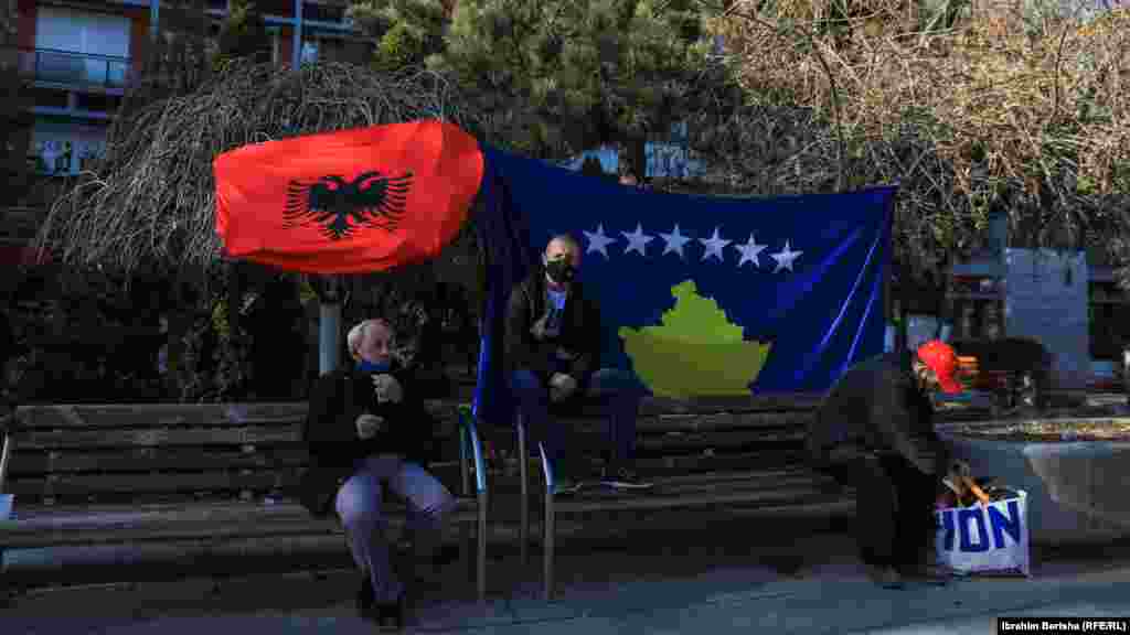 Disa qytetarë janë ulur në sheshin &ldquo;Nëna Terezë&rdquo;, prapa tyre qëndron flamuri i Kosovës dhe ai i Shqipërisë.