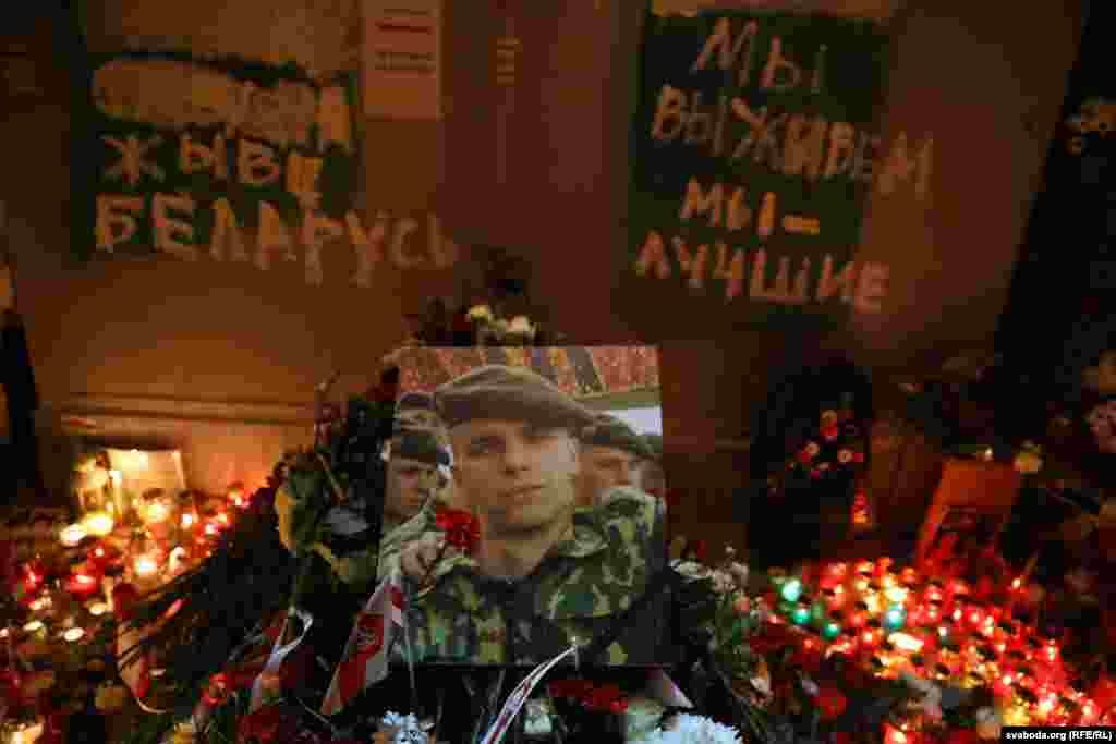 A photograph of Raman Bandarenka is surrounded with candles and flowers brought by the mourners. The sign on the left says: &quot;[Long] live Belarus,&quot; and the one on the right says: &quot;We will survive, we are the best.&quot;