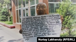 Зейнеткер Райса Дүйсенбаева полиция ғимаратының алдында плакат ұстап тұр. Алматы, 26 шілде 2018 жыл